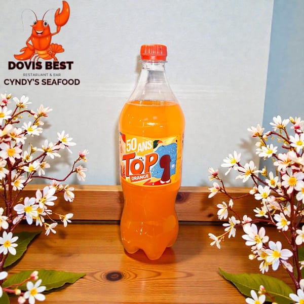 A bottle of orange soda sits on a wooden surface, framed by white flowers and green leaves.