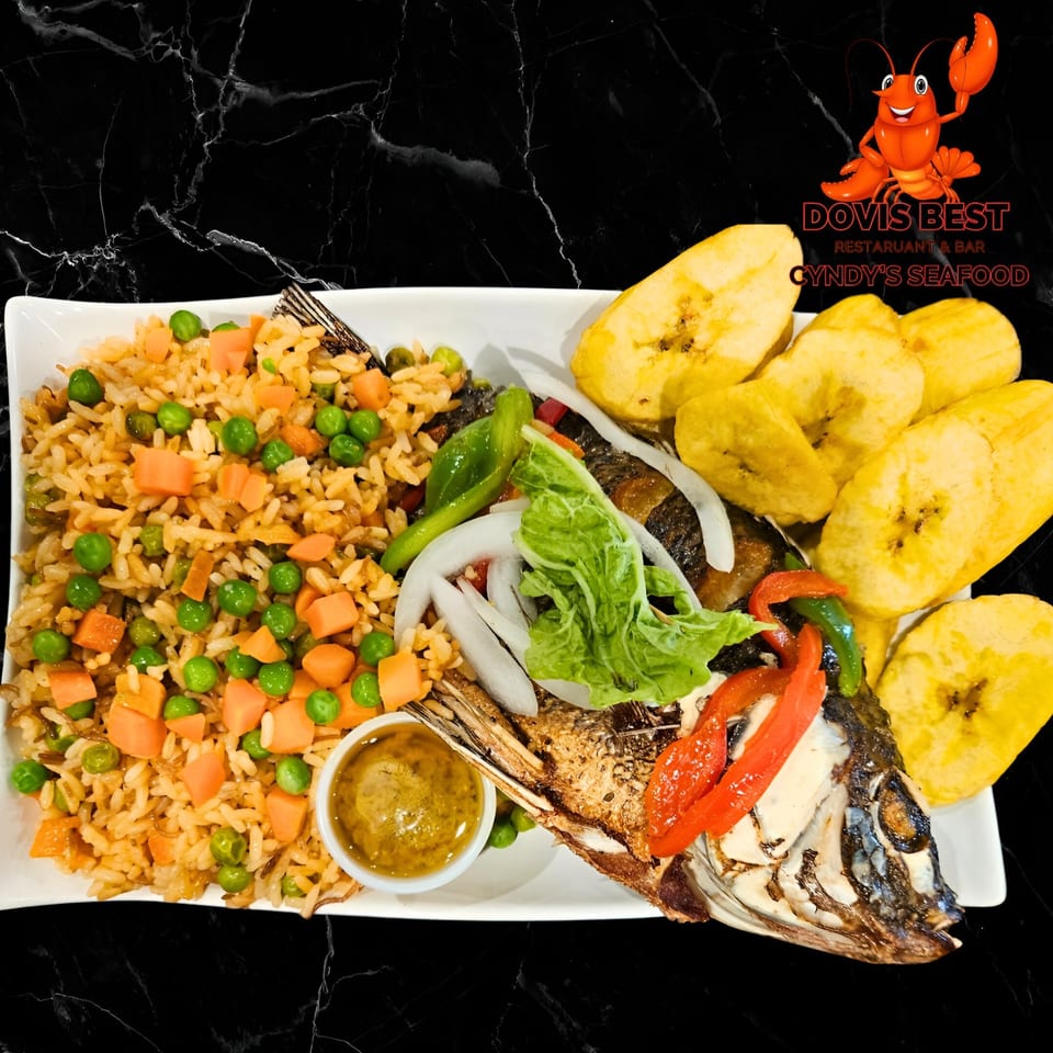 A whole fried fish with rice, peas, carrots, plantains, and sauce on a white plate.
